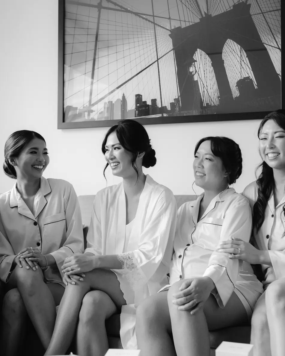 The bride and her bridesmaids in matching white pajamas, laughing together on a sofa. Black and white.