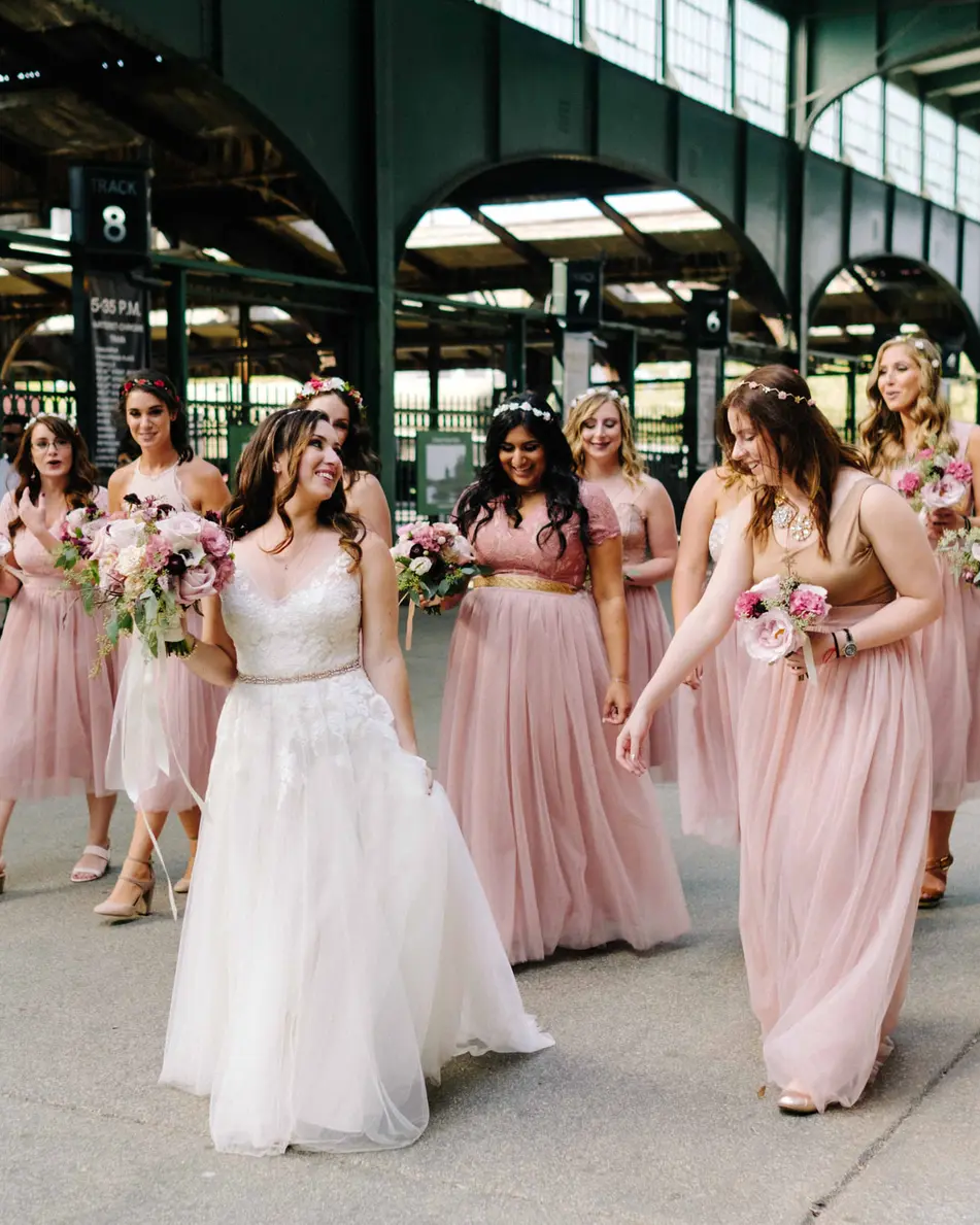 The bride walks with her bridesmaids in blush beneath the green steel canopy of the old railroad terminal.