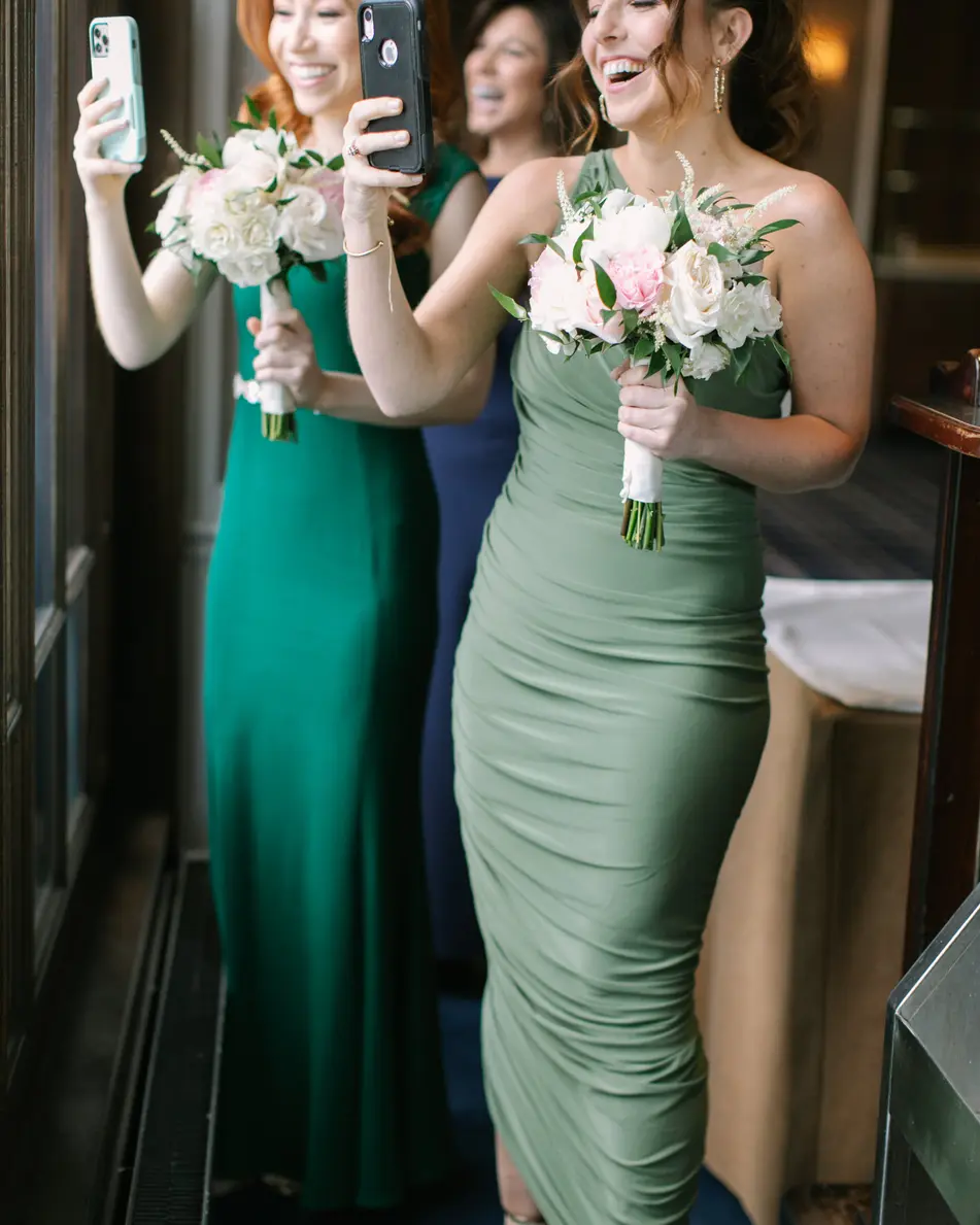 Two bridesmaids in green hold up their phones and bouquets, smiling as they watch from a window.