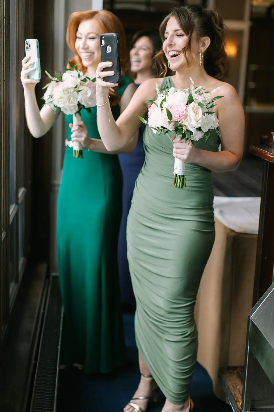 Two bridesmaids in green hold up their phones and bouquets, smiling as they watch from a window.