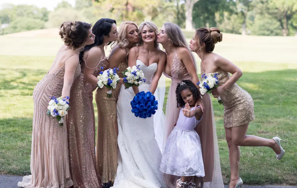 The bridesmaids in blush and gold sequins kiss the bride on the cheek, a flower girl beside them.