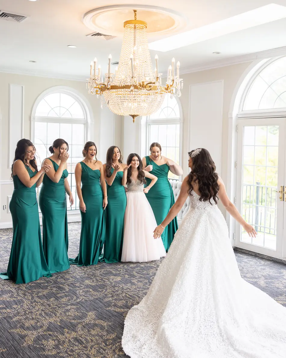 In a room of tall arched windows, bridesmaids in emerald gowns clasp their hands as the bride turns to face them.