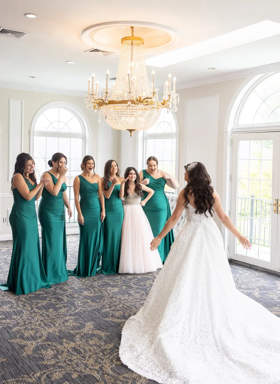 In a room of tall arched windows, bridesmaids in emerald gowns clasp their hands as the bride turns to face them.