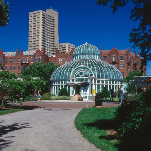 The glass Palm House at Brooklyn Botanic Garden in summer, framed by trees and lawn under a clear blue sky.