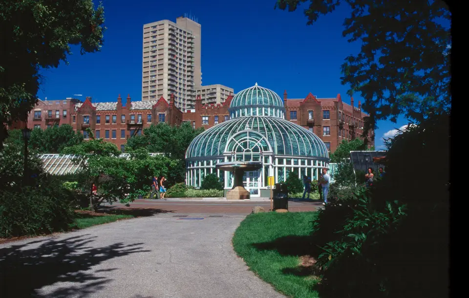 The glass Palm House at Brooklyn Botanic Garden in summer, framed by trees and lawn under a clear blue sky.