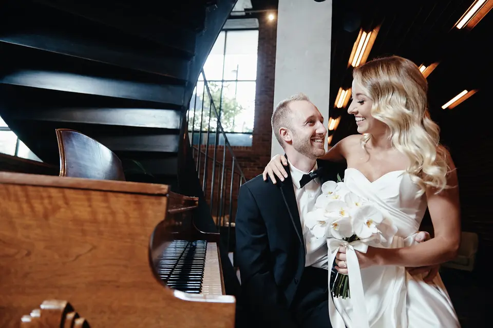 The couple embrace beside an old piano, the bride holding white orchids.