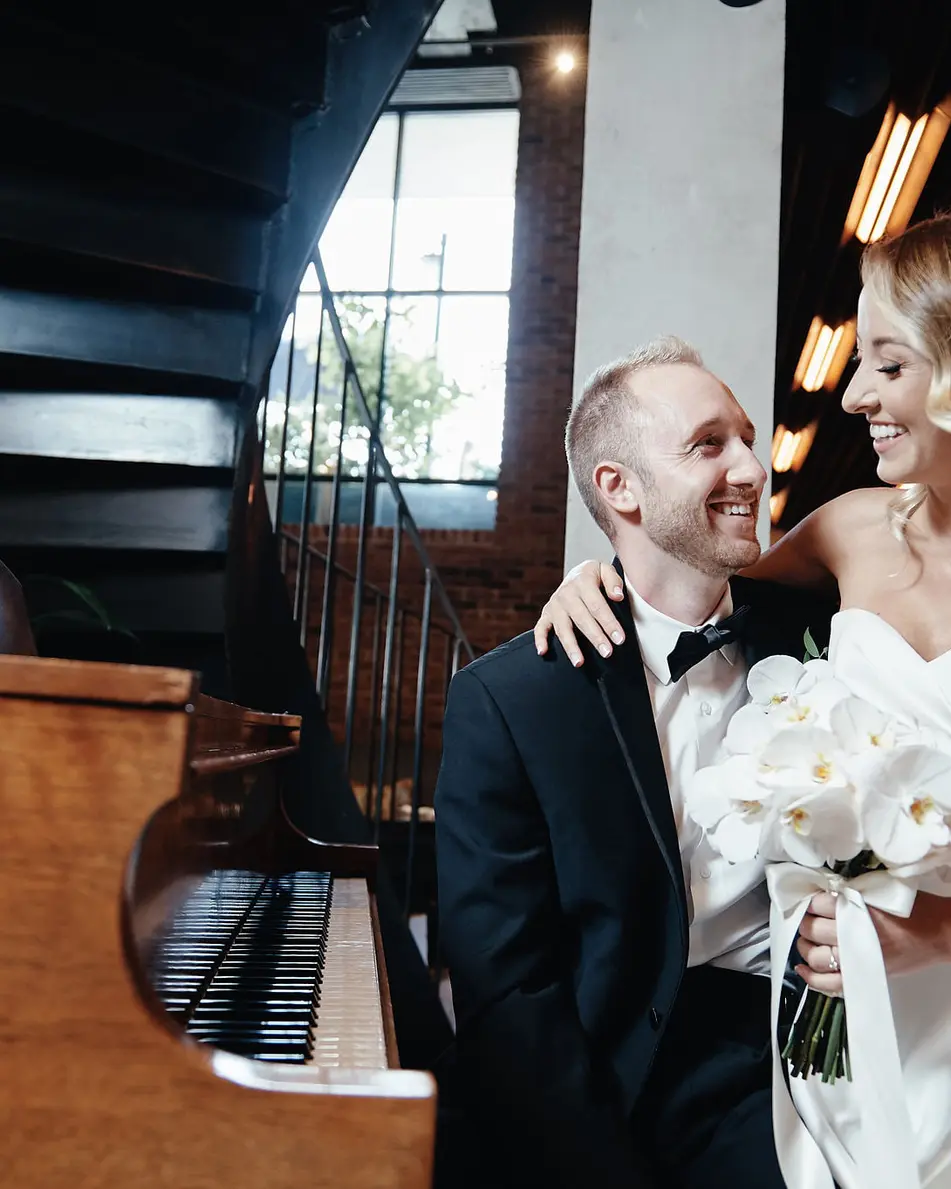 The couple embrace beside an old piano, the bride holding white orchids.