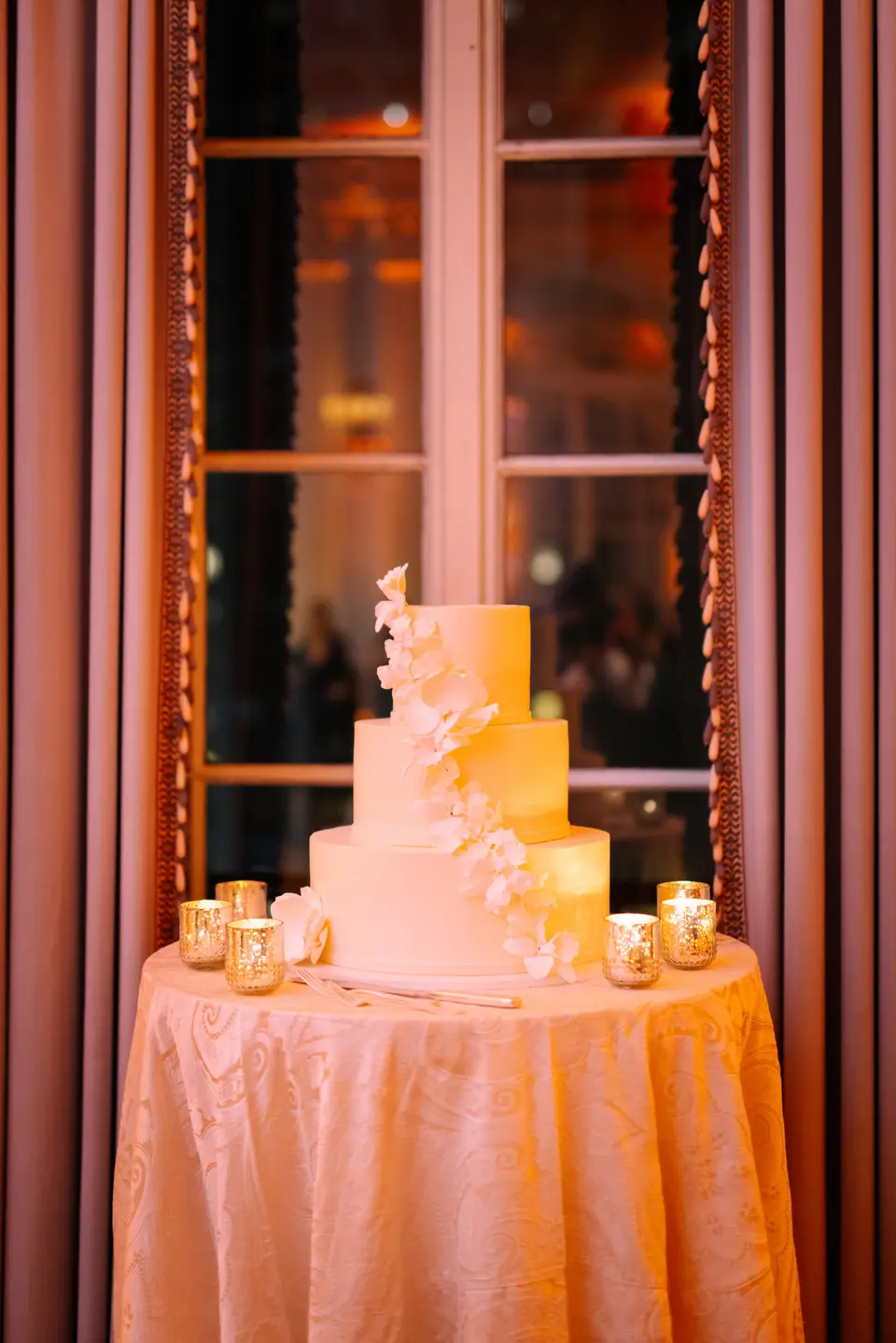 A tall white cake with sugar flowers on a round table, candles around it, before a tall window at night.