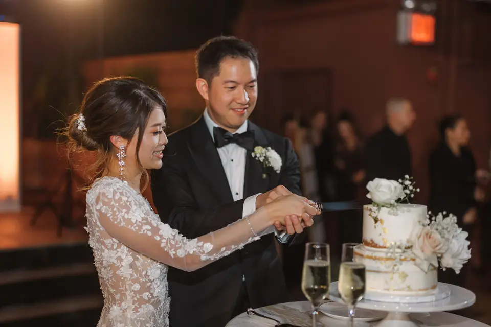 The couple cut a small white and gold cake with flowers, glasses of champagne beside it.