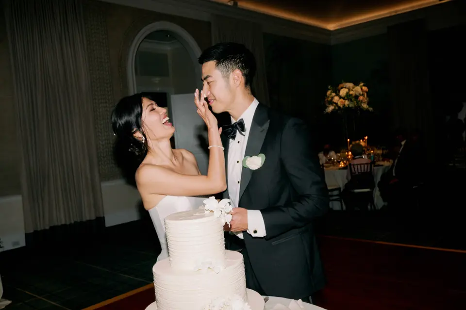 The bride wipes a little cake from the groom's cheek beside a tall white cake, both laughing.
