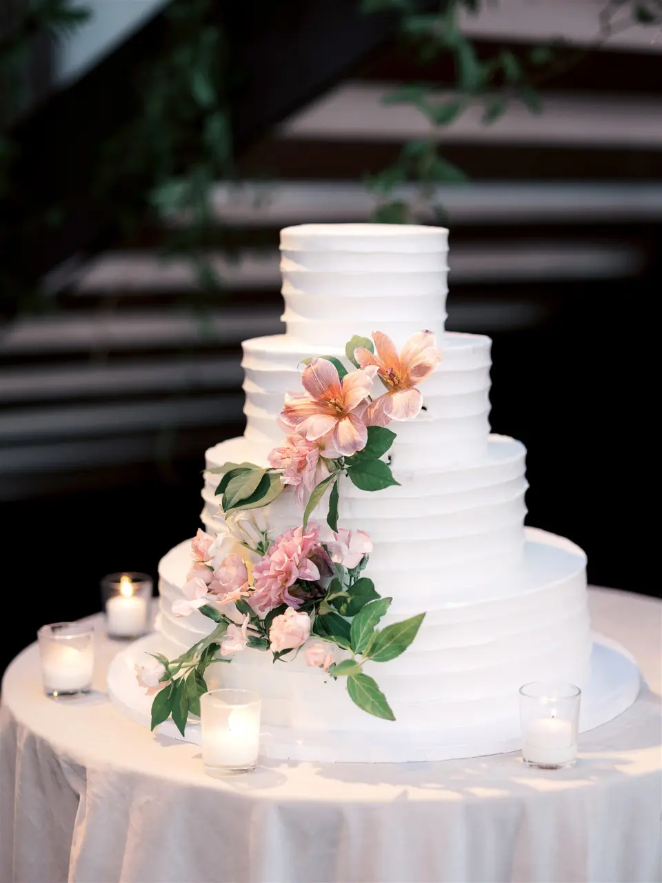 A four-tier white cake with pink lilies and greenery on a round table lit by candles.