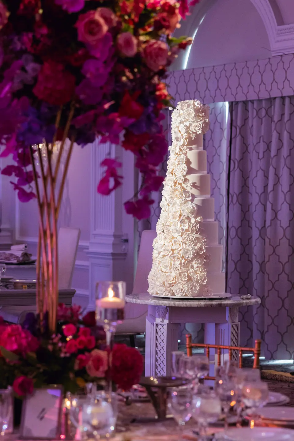 A tall white cake beside a pillar of pink and red flowers, candles and glassware on the tables around it.