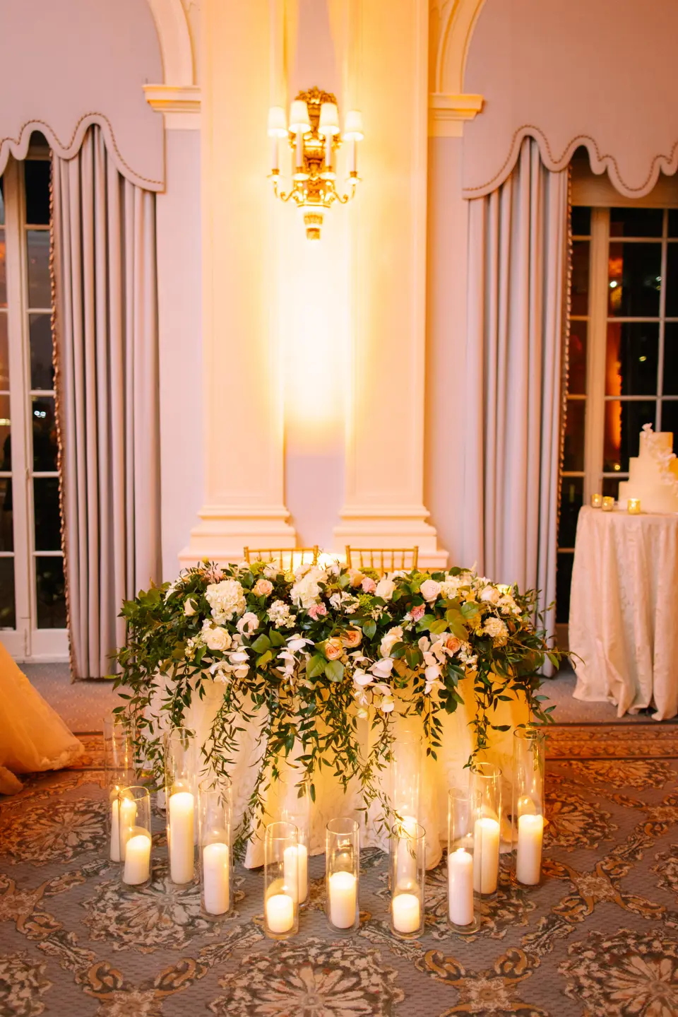 A low bank of white flowers and greenery surrounded by candles glows at the foot of a pale column.