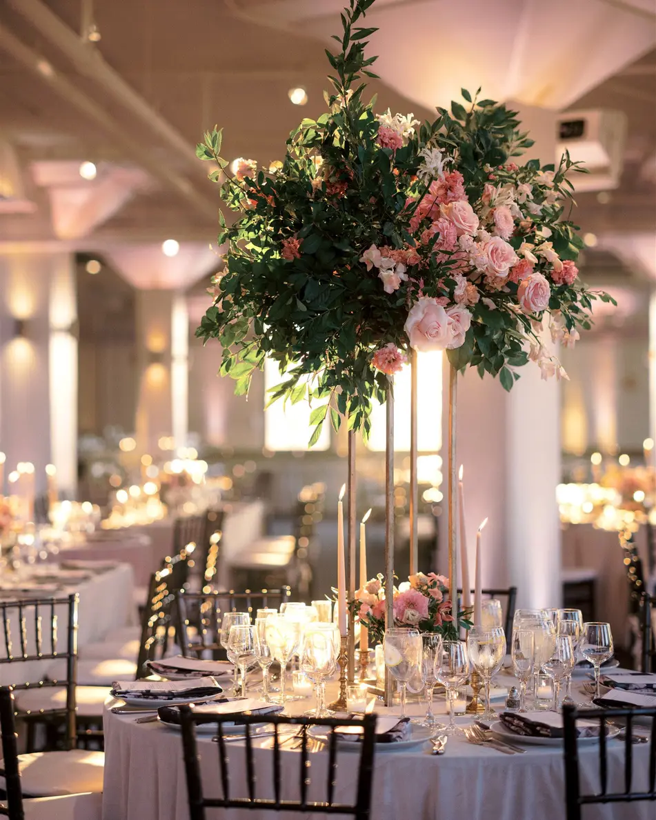 A tall arrangement of pink roses and greenery on a gold stand rises above a round table set with candles and glassware.