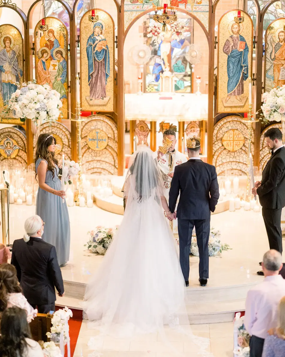 The couple stand before a gilded wall of painted icons at the altar of the church, candles and flowers around them.