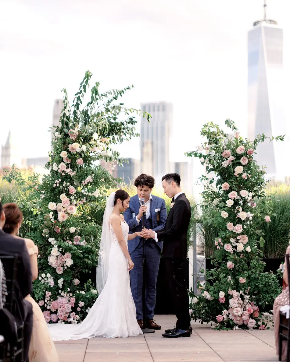Beneath an arch of pink roses and greenery, the couple stand before their officiant, One World Trade Center behind them.