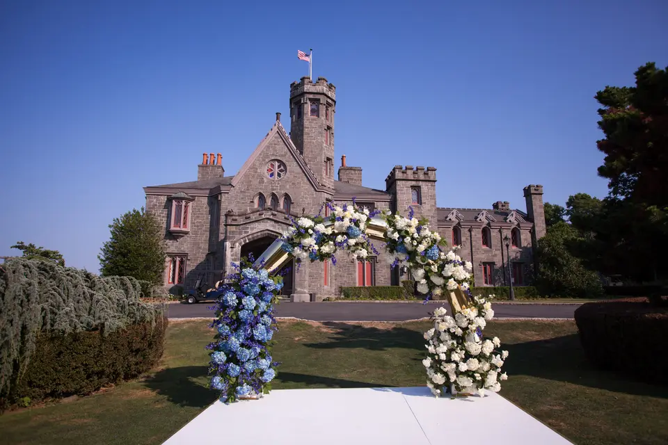 A circular arch of blue and white flowers on a white platform on the lawn, the castle behind under a clear blue sky.