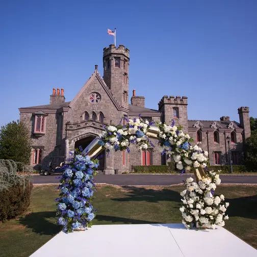 A circular arch of blue and white flowers on the lawn at Whitby Castle, the castle behind under a clear blue sky.