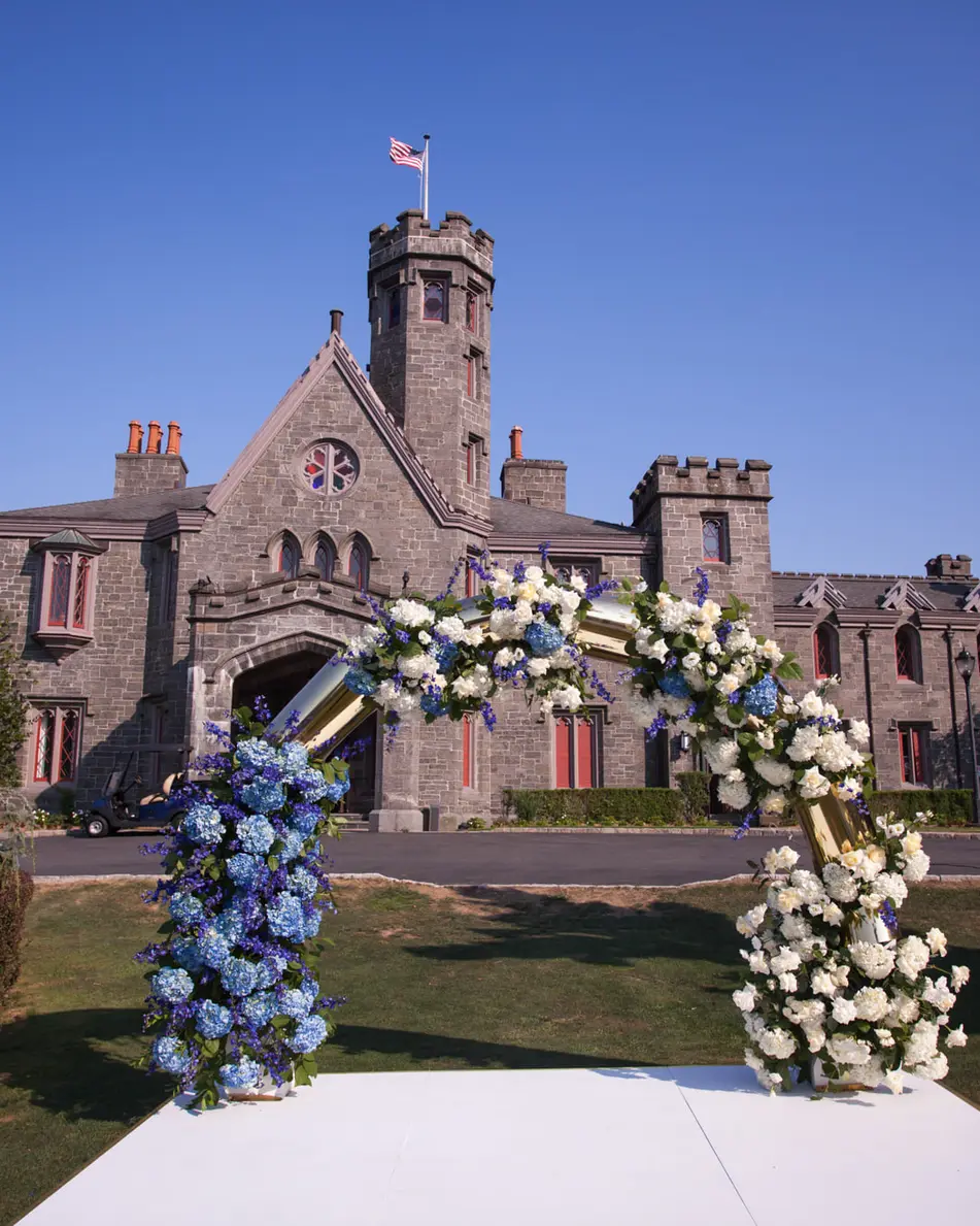 A circular arch of blue and white flowers on a white platform on the lawn, the castle behind under a clear blue sky.
