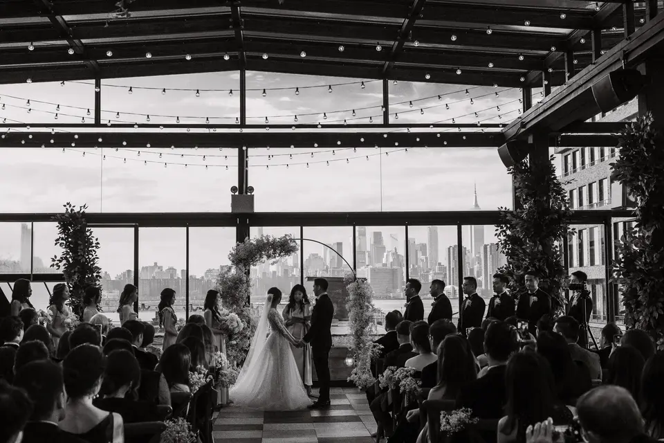 The ceremony seen from the back of the room, guests silhouetted against the tall windows and the skyline. Black and white.