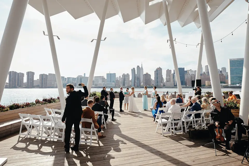 Guests gather beneath the white canopy on the waterfront, facing the Manhattan skyline across the river.