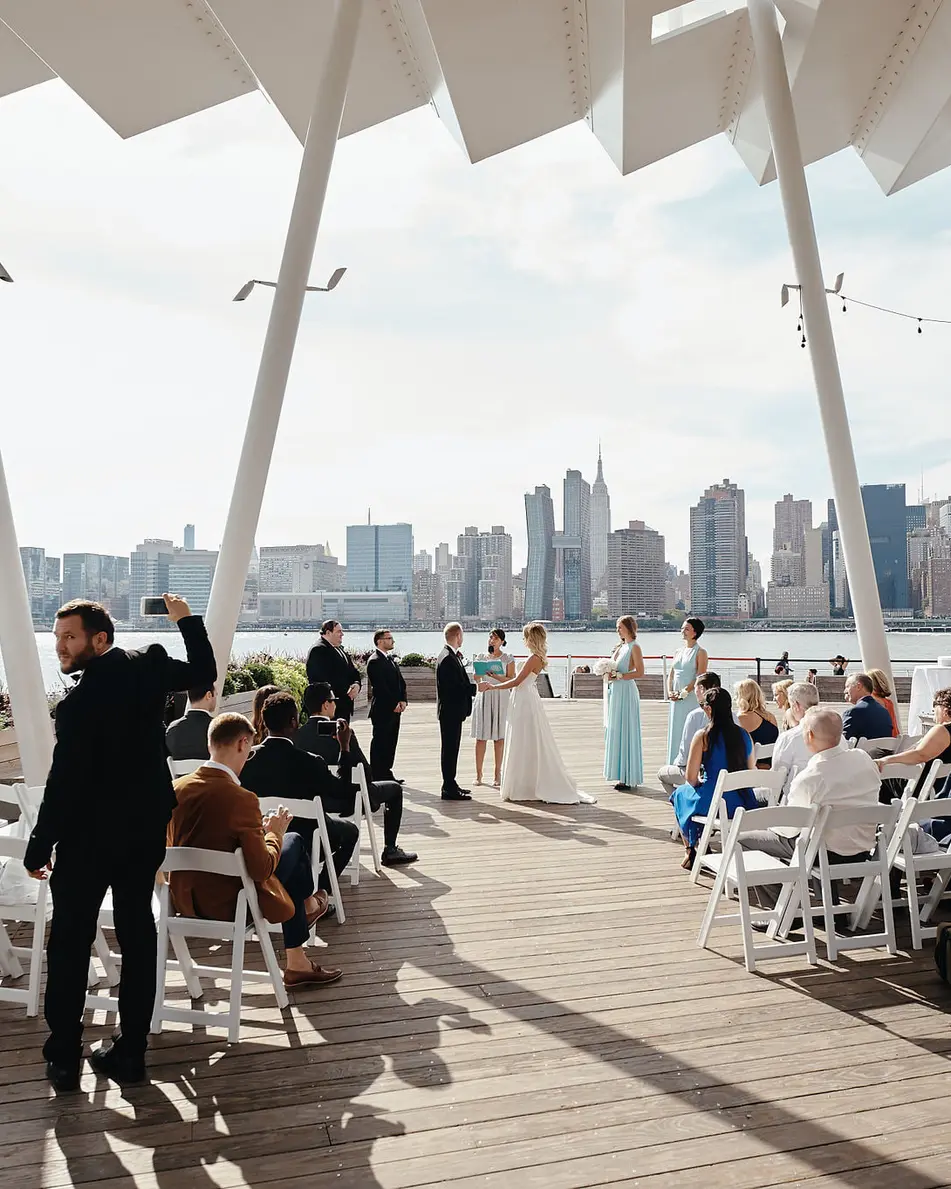 Guests gather beneath the white canopy on the waterfront, facing the Manhattan skyline across the river.