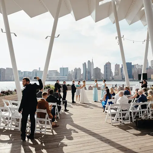 Guests gather beneath a white canopy on the Long Island City waterfront, facing the Manhattan skyline across the East River.