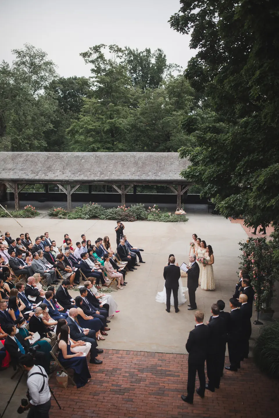 Seen from above, guests fill rows of chairs in the stone courtyard as the couple stand before the officiant, trees all around.