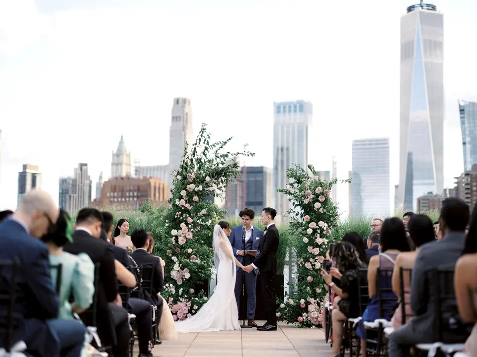Guests watch from both sides of the aisle as the couple stand beneath the rose arches, the skyline beyond.