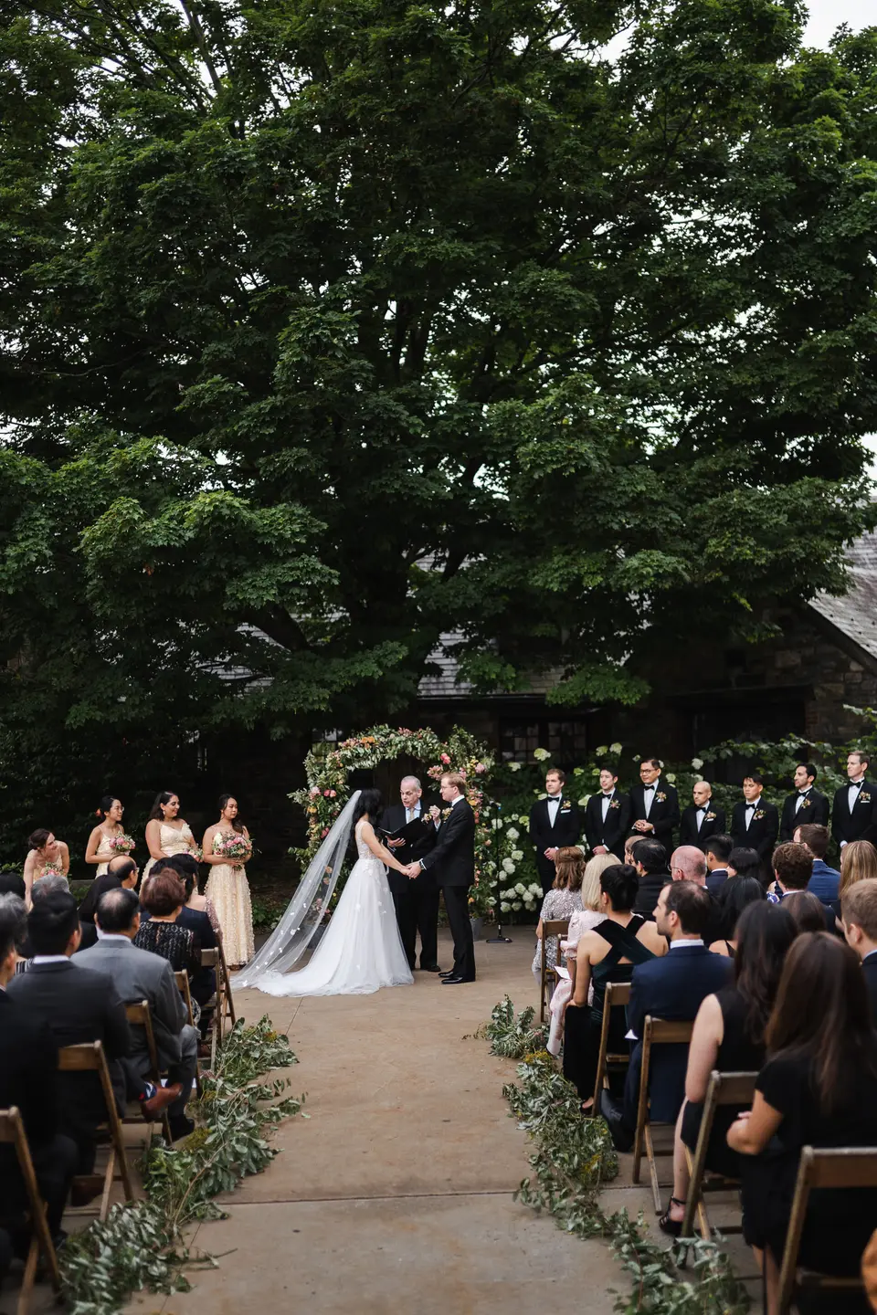Guests seated on either side of a gravel aisle lined with greenery, the couple at the far end beneath the trees.