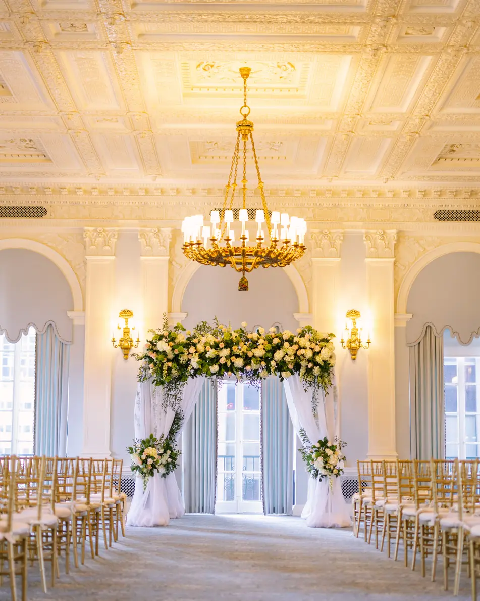 A pale ballroom with a crystal chandelier, rows of gold chairs facing a chuppah of white and green flowers hung with white drapes.