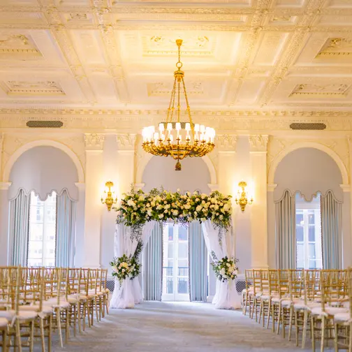 A pale ballroom at The Yale Club with a crystal chandelier, rows of gold chairs facing a chuppah of white and green flowers hung with white drapes.
