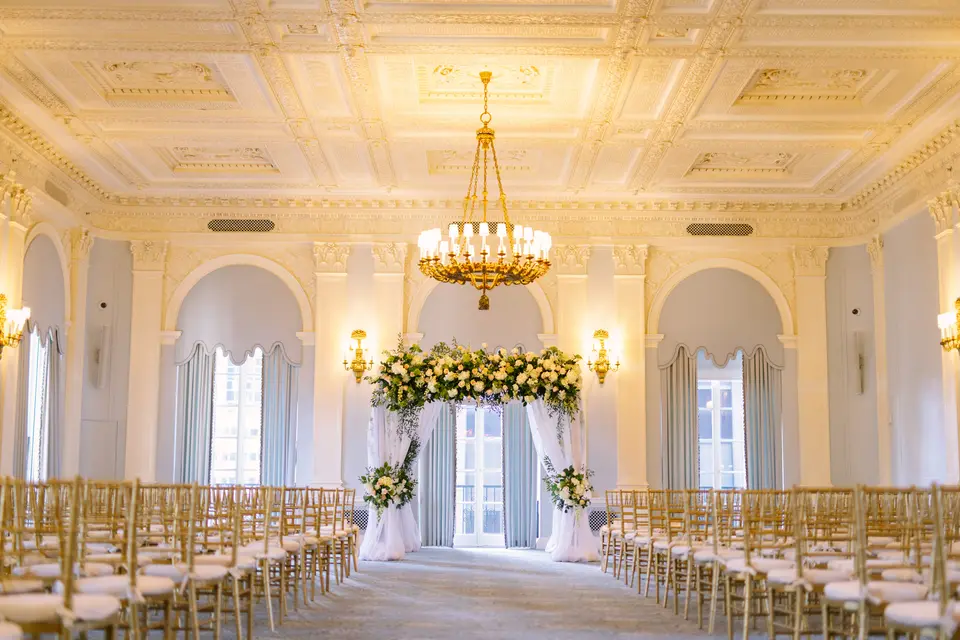 A pale ballroom with a crystal chandelier, rows of gold chairs facing a chuppah of white and green flowers hung with white drapes.