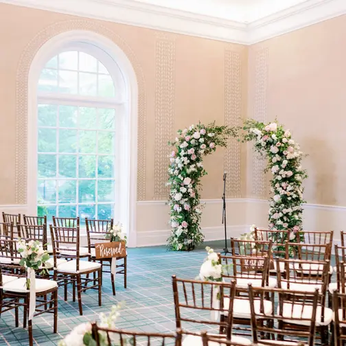 A room at The New York Botanical Garden with arched windows, rows of wooden chairs facing an arch of white and blush flowers and greenery.