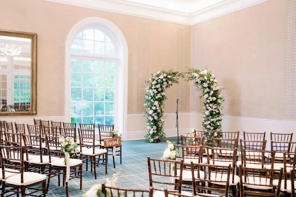 A room with arched windows, rows of wooden chairs facing an arch of white and blush flowers and greenery.