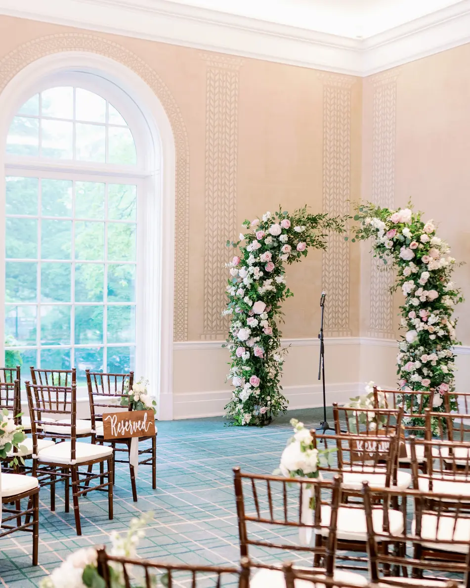 A room with arched windows, rows of wooden chairs facing an arch of white and blush flowers and greenery.