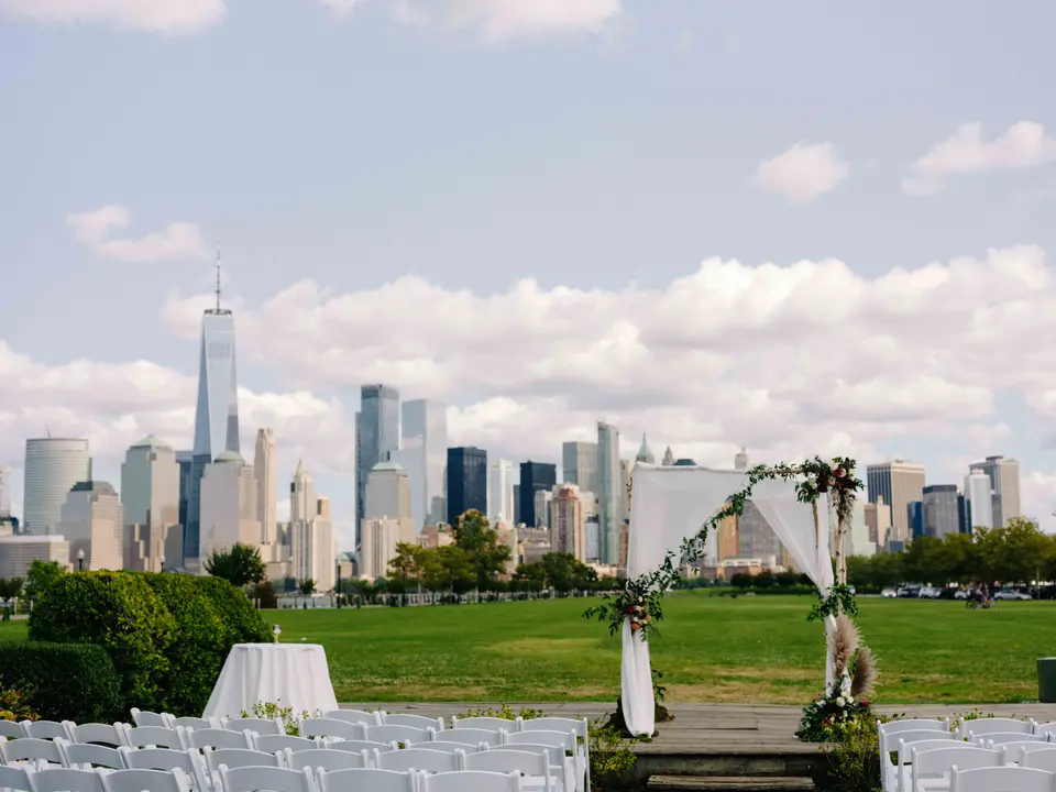 On the lawn, rows of white chairs face a wooden arch draped with white fabric and flowers, the Manhattan skyline across the river.