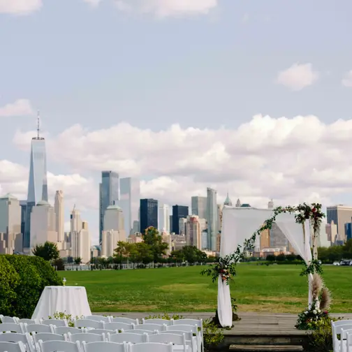 On the lawn at Liberty House, rows of white chairs face a wooden arch draped with white fabric and flowers, the Manhattan skyline across the river.
