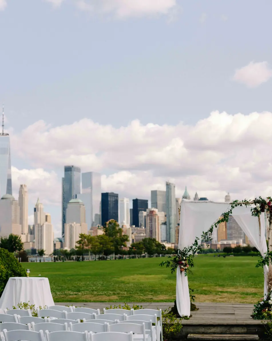 On the lawn, rows of white chairs face a wooden arch draped with white fabric and flowers, the Manhattan skyline across the river.