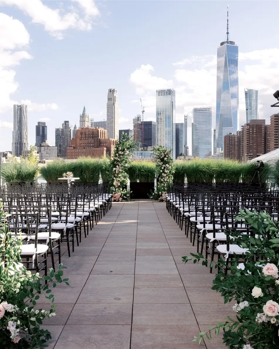 Rows of black chairs on the rooftop face two arches of roses, the towers of Lower Manhattan beyond them under a blue sky.