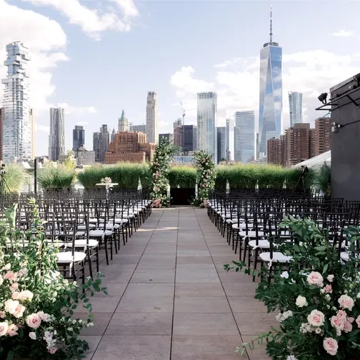 Rows of black chairs on the roof at Tribeca Rooftop face two arches of roses, the towers of Lower Manhattan beyond them under a blue sky.