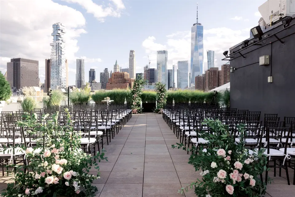 Rows of black chairs on the roof at Tribeca Rooftop face two arches of roses, the towers of Lower Manhattan beyond.