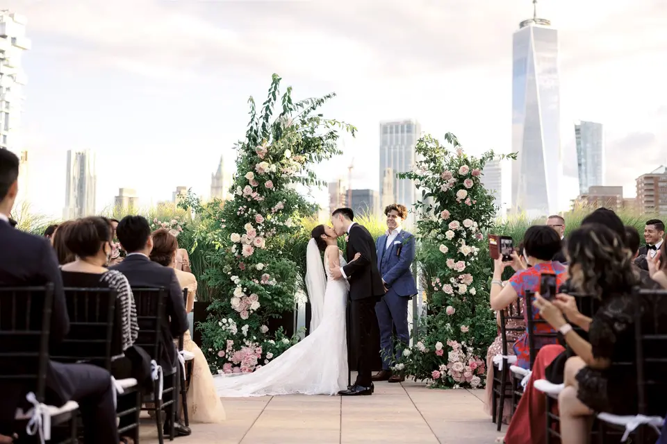 On the roof at Tribeca Rooftop, the couple kiss between two tall arches of pink roses and greenery, guests seated on either side and One World Trade Center rising behind them.