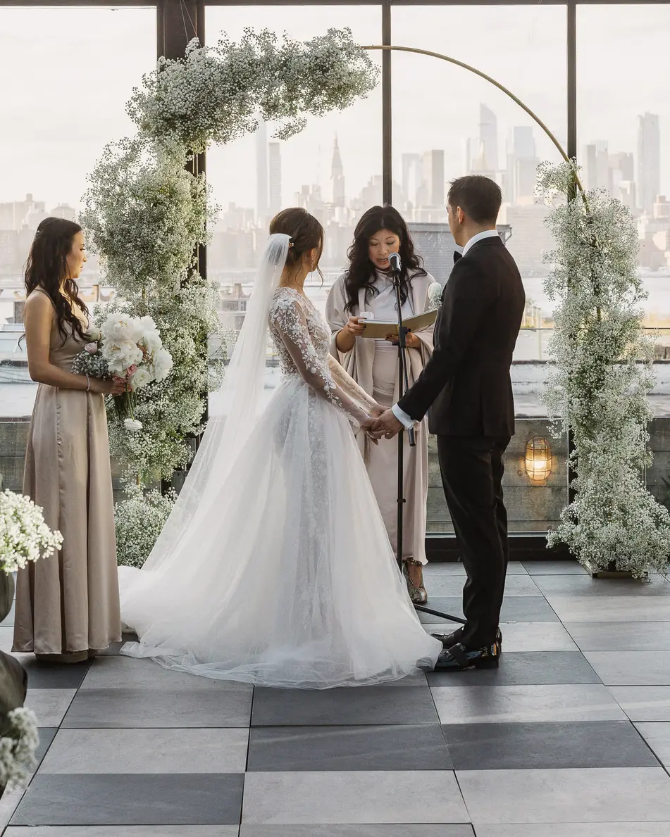 In a glass-walled room with a checkerboard floor, the couple hold hands before the officiant beneath an arch of white flowers, the skyline beyond the windows.