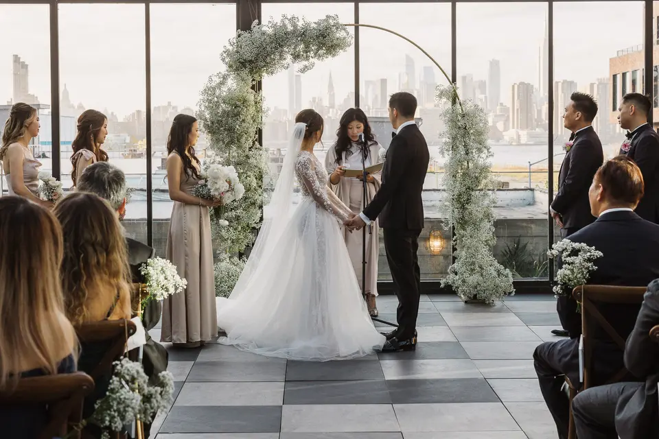 In a glass-walled room at 74 Wythe, the couple hold hands before the officiant beneath an arch of white flowers, the Manhattan skyline beyond the windows.