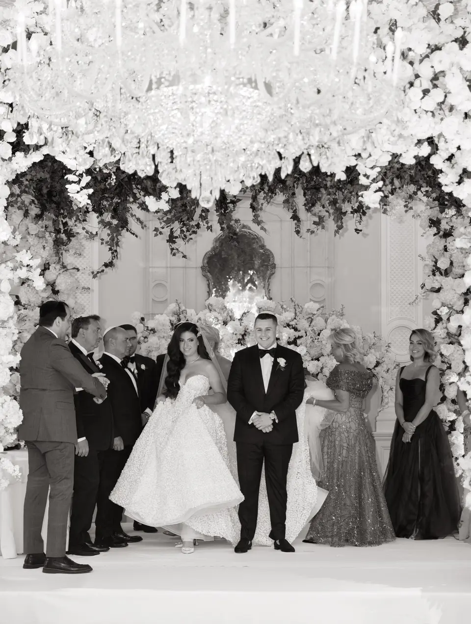 The couple stand with their families beneath a canopy of white flowers and crystal. Black and white.