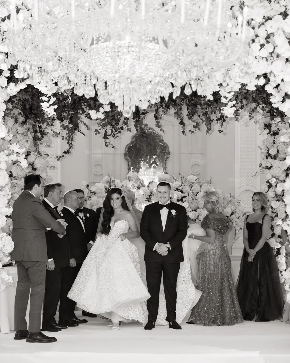 The couple stand with their families beneath a canopy of white flowers and crystal. Black and white.