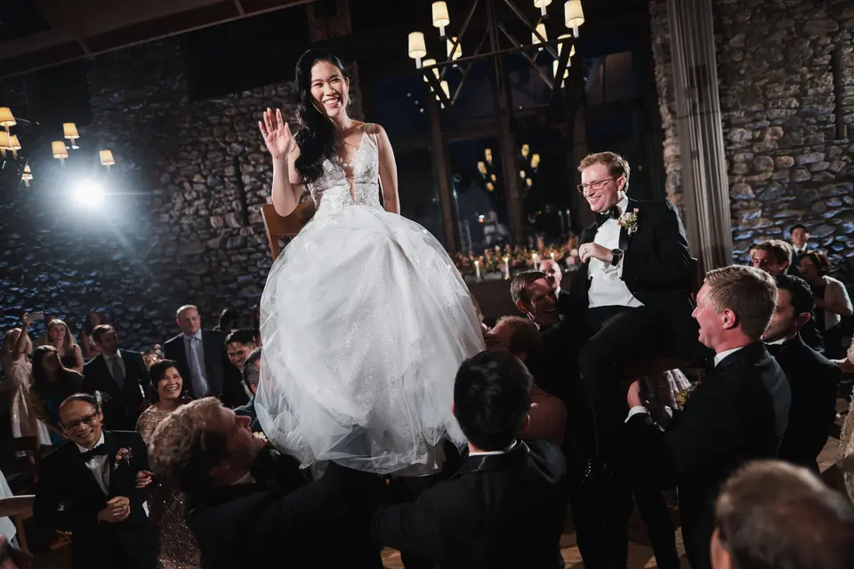 The bride waves from a chair lifted high above the guests, the groom lifted beside her.