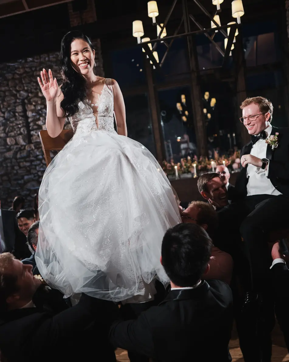 The bride waves from a chair lifted high above the guests, the groom lifted beside her.