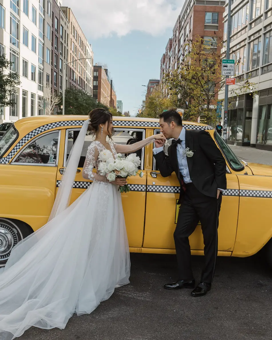 On a Williamsburg street, the groom leans in at the open door of a yellow checker cab and kisses his bride's hand, her veil and train spread across the road.